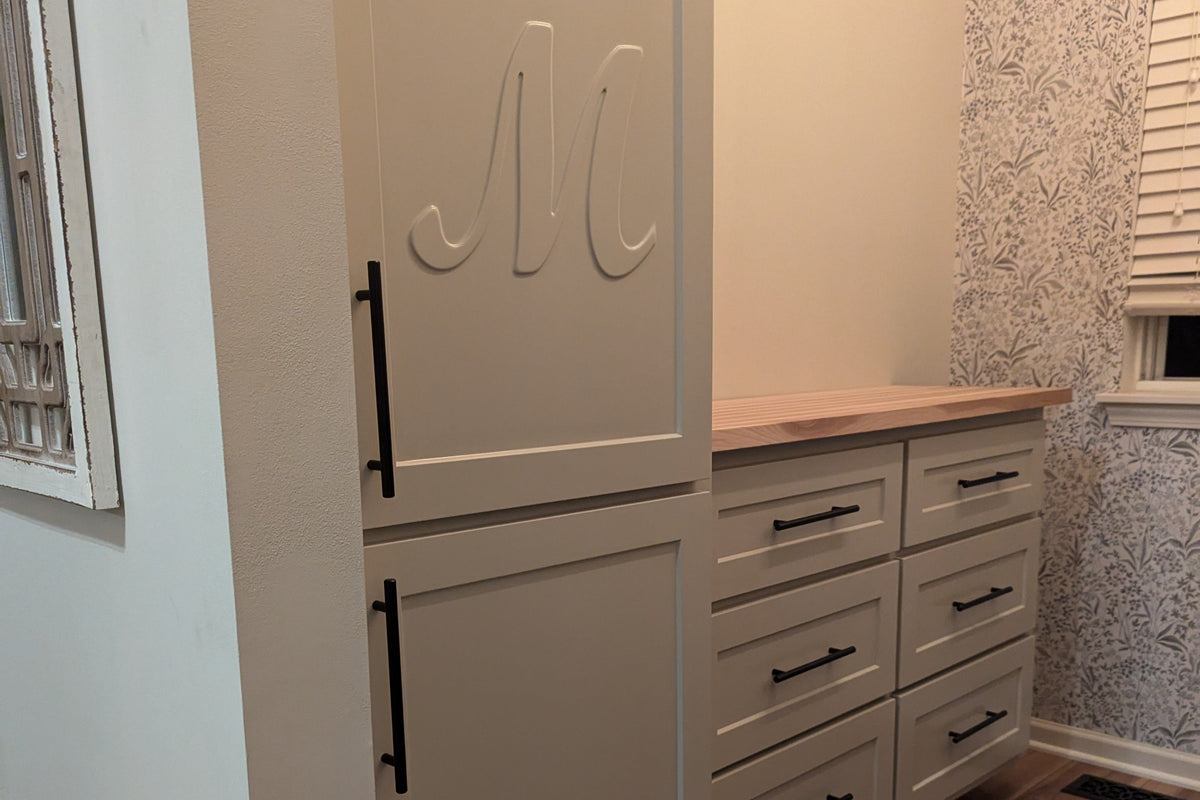 A tall, light beige entryway cabinet with black handles and a matching drawer unit with a light wood top, installed in a home hallway with wood floors and patterned wallpaper.